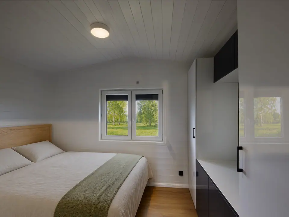 Schlafzimmer des Modulhauses TINY-MAX 35-1 mit Doppelbett, hellem Holzkopfteil und weißem Einbauschrank, am Fenster Blick auf Birken und Wiese. Visualisierung
