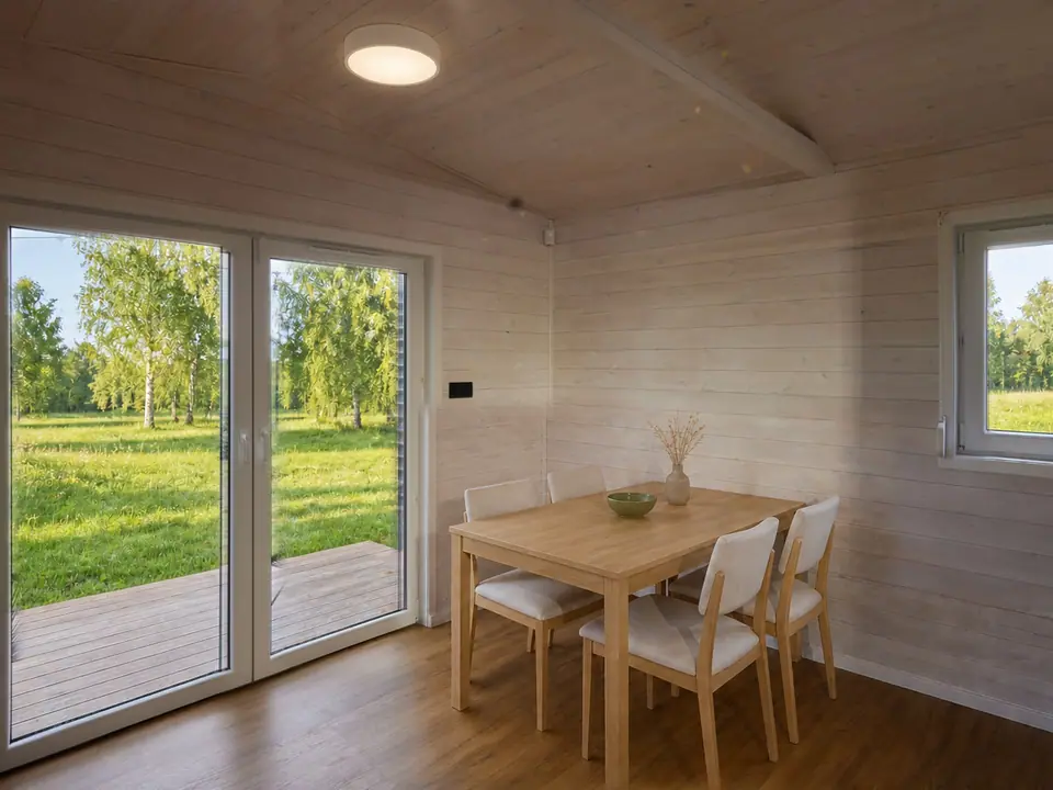 Heller Wohn- und Essbereich des Modulhauses TINY-MAX 35-1 mit Esstisch aus Eiche und Polsterstühlen, dahinter eine bodentiefe zweiflügelige Terrassentür mit Blick auf eine Wiese mit Birken. Wände und Decke in heller Holzschalung. Visualisierung