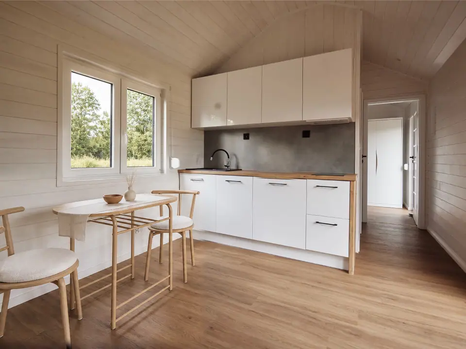 Offener Wohnbereich mit weißer Küchenzeile im Modulhaus TINY-MAX 35-2, davor ein ovaler Esstisch mit Holzstühlen, links ein Fenster mit Blick ins Grüne. Wände und die gewölbte Decke in heller Holzschalung. Visualisierung
