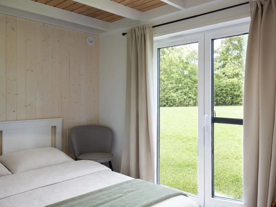 Eines der drei Schlafzimmer im Modulhaus TINY-MAX 47 mit Doppelbett, weißem Holzbettgestell und grauem Polsterstuhl, einer Wandverkleidung aus hellem Fichtenholz und einer bodentiefen Terrassentür mit hellen Vorhängen, dahinter Rasen und Hecke. Visualisierung