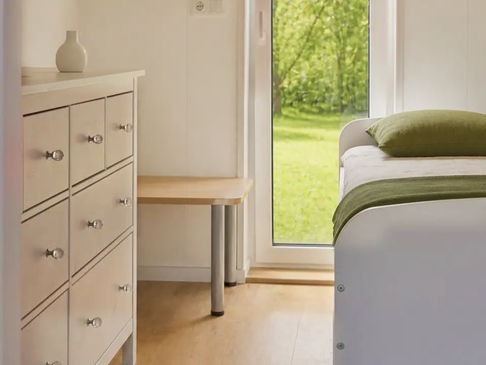 Schlafzimmer des Modulhauses TINY-MAX 59 mit heller Kommode, Bett und einer Fenstertür mit Blick auf die Wiese. Visualisierung