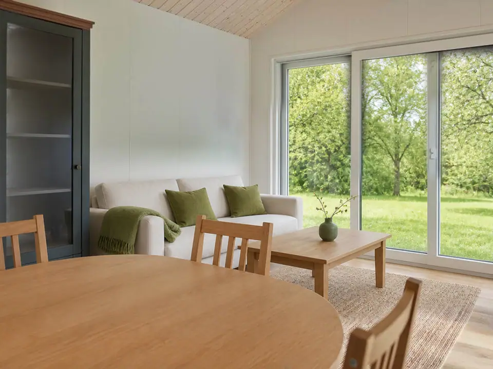 Wohnbereich des Modulhauses TINY-MAX 59 mit hellem Sofa, Couchtisch aus Eiche und großen Schiebefenstern zum Garten, im Vordergrund der runde Esstisch. Visualisierung